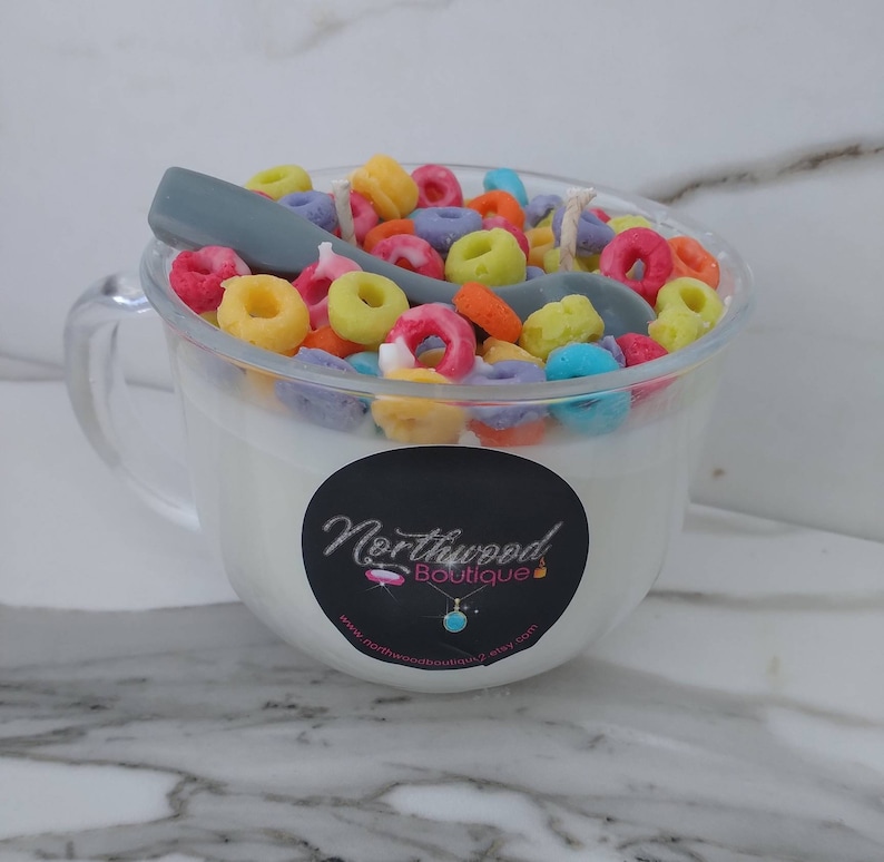 May include: A clear glass mug candle filled with white wax and colorful cereal pieces. A gray spoon rests on top. A black sticker with the text "Northwood Boutique" is on the front of the mug.