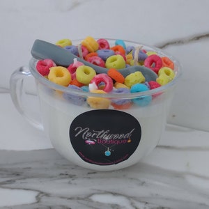 May include: A clear glass mug-shaped candle filled with white wax, topped with colorful cereal pieces and a gray spoon. A black sticker with white text and a turquoise pendant is on the front of the mug.