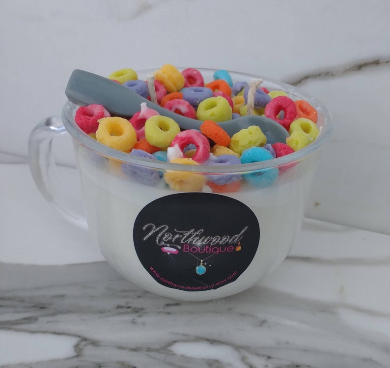 May include: A clear glass mug-shaped candle filled with white wax, topped with colorful cereal pieces and a gray spoon. A black sticker with white text and a turquoise pendant is on the front of the mug.