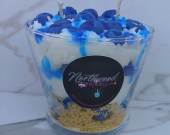 May include: A clear glass candle holder filled with layers of white, blue, and tan wax, topped with blue wax blueberries. The candle has two wicks and a black label with the text "Northwood Boutique".
