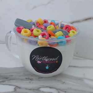May include: A clear glass mug-shaped candle filled with white wax, topped with colorful cereal pieces and a gray spoon. A black sticker with the words "Northwood Boutique" is on the front of the mug.