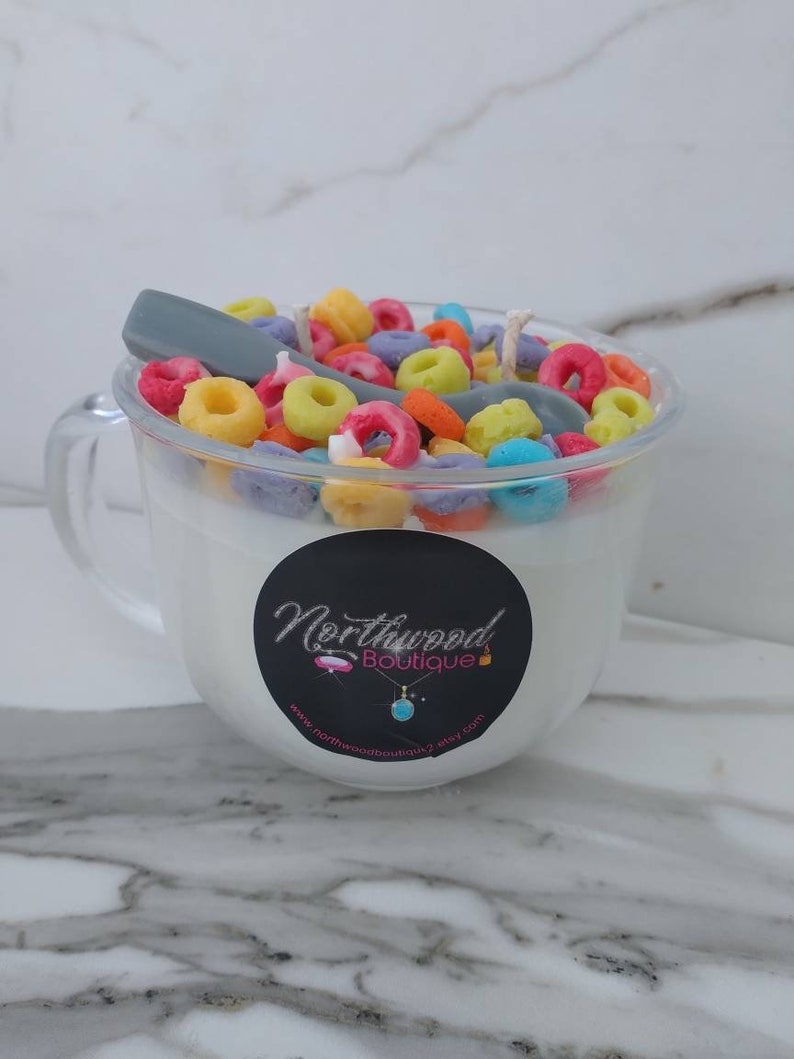 May include: A clear glass mug-shaped candle filled with white wax, topped with colorful cereal pieces and a gray spoon. A black sticker with the words "Northwood Boutique" is on the front of the mug.