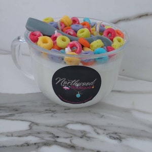 May include: A clear glass mug-shaped candle filled with white wax and colorful, cereal-shaped wax pieces. A gray spoon-shaped wax piece rests on top. The candle has a black label with the text "Northwood Boutique".