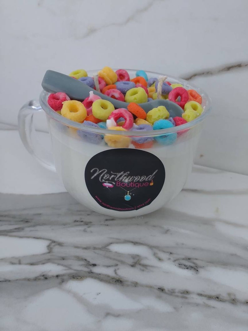 May include: A clear glass mug-shaped candle filled with white wax and colorful, cereal-shaped wax pieces. A gray spoon-shaped wax piece rests on top. The candle has a black label with the text "Northwood Boutique".