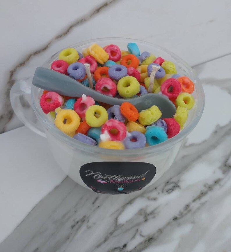 May include: A novelty candle shaped like a bowl of colorful cereal with a gray spoon. The candle is in a clear glass cup with a handle. The candle has a white base and is topped with colorful, ring-shaped wax pieces.