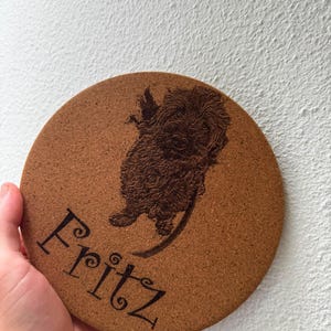 Può includere: Un sottobicchiere rotondo in sughero marrone chiaro con un'illustrazione incisa al laser in marrone scuro di un cane dal pelo arruffato e il nome Fritz scritto in un carattere corsivo stilizzato. Il sottobicchiere ha una superficie granulosa.