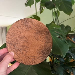 Puede incluir: Un posavasos circular de corcho marrón con un patrón grabado a láser de varias hojas de monstera. La superficie del posavasos tiene una textura natural y granulada, y los diseños de las hojas están detallados con líneas finas y recortes.