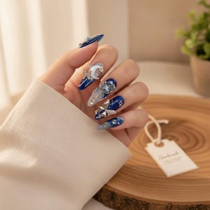May include: Long almond-shaped nails featuring a deep blue base with intricate silver butterfly charms, a crescent moon accent, sparkling rhinestones, and delicate star-shaped embellishments. The design incorporates a mix of glossy and glittery finishes.
