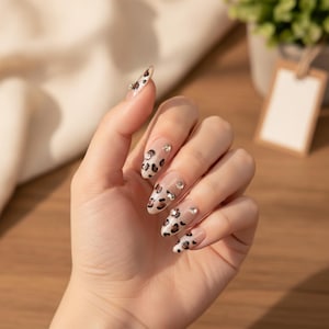 Peut inclure: Ongles press-on en forme d'amande avec une base transparente ornée d'un motif léopard marron et noir. Chaque ongle est agrémenté de petits strass ronds et transparents placés près de la cuticule et sur les côtés du motif.
