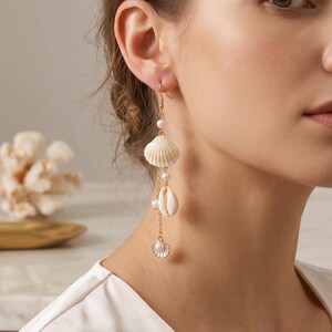 Peut inclure: Boucles d'oreilles pendantes composées de chaînes dorées ornées de petits coquillages blancs, incluant des formes de coquilles Saint-Jacques et de cauris, entremêlés de minuscules perles blanches. Elles sont suspendues à des crochets dorés.