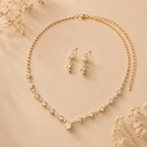 May include: A gold-toned necklace and matching pair of drop earrings, each featuring a series of clear, round-cut stones set in a delicate metal chain. The necklace includes an adjustable chain with a lobster clasp and a central stone focal point.