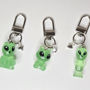 Könnte beinhalten: Drei Schlüsselanhänger mit durchscheinenden hellgrünen Alien-Figuren mit großen schwarzen Augen. Jedes Alien ist an einem silberfarbenen Metallkarabiner und einem kleinen silbernen Sternanhänger befestigt. Die Figuren haben unterschiedliche Posen.