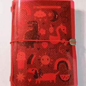 Può includere: Una copertina per taccuino rossa, traslucida e glitterata, caratterizzata da un motivo ripetuto di icone rosso scuro che includono un dinosauro, un fenicottero, un bassotto, un gelato, un sole, un arcobaleno, una fetta d'anguria, una nuvola, una luna e una matita.