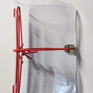 May include: A clear, flexible plastic cover featuring a bright red elastic cord system. The cord is threaded through vertical slots on the left side and secured with a small metal clasp on the right, creating a tension-based closure mechanism.