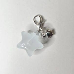 May include: A silver-toned metal lobster clasp charm featuring a smooth, opaque white star-shaped pendant and a smaller, solid silver-toned star charm attached to the same jump ring. The design is simple, with a polished finish on the metal components.