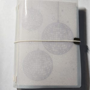 Puede incluir: Una cubierta de cuaderno translúcida y brillante con un diseño impreso de tres bolas de discoteca. La cubierta se mantiene cerrada mediante una fina banda elástica blanca con un pequeño clip metálico en el lateral, asegurando las páginas.