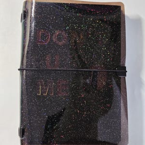 May include: A black glittery notebook cover with a translucent finish and a black elastic band closure. The text "DON'T U ME" is printed in a metallic font on the front cover, which features a multi-colored holographic sparkle effect throughout.