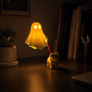Op de afbeelding: Een kleine, gloeiende, gele lamp in de vorm van een geest, bevestigd met een rode riem aan een beeldje van een lichtgekleurde puppy. De geestlamp geeft een warm, zacht licht en verlicht het puppybeeldje op een donker oppervlak.