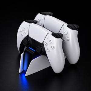 Puede incluir: Dos mandos de juego inalámbricos blancos y negros colocados en una estación de carga blanca. Los mandos cuentan con una cruceta, dos joysticks analógicos y cuatro botones de acción con símbolos de triángulo, círculo, cruz y cuadrado. Una luz azul brilla.