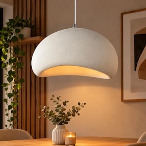Puede incluir: Una lámpara colgante en forma de cúpula con un acabado mate texturizado en color blanco roto. La lámpara presenta una abertura ancha y curva en la parte inferior que emite un brillo cálido y suave desde una fuente de luz interna, suspendida por un cable blanco.