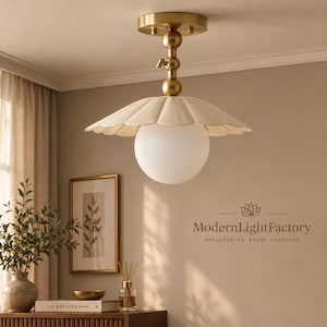 May include: A ceiling light fixture featuring a brass finish mount with a central stem and a scalloped, cream-colored metal shade. A spherical white glass globe hangs below the shade. The text ModernLightFactory Handcrafted Brass Lighting is visible.