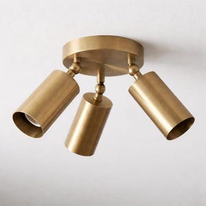 May include: A three-light ceiling fixture with a brushed brass finish. The circular base supports three adjustable cylindrical spotlights, each connected by a swivel joint, allowing for directional lighting from the metallic lamp heads.