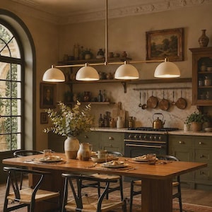 May include: A linear pendant light fixture featuring four matte white dome-shaped shades suspended from a long, thin brass-toned metal rod. The fixture is positioned horizontally above a wooden dining table in a kitchen setting.