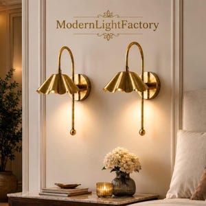 以下が含まれることがあります： ModernLightFactoryという文字が記された壁に取り付けられた、真鍮製の壁掛け照明2つ。波打つベル型のシェードと曲線的なアームが特徴で、下向きの光源と小さな球体で終わる細い垂直のロッドを備えています。