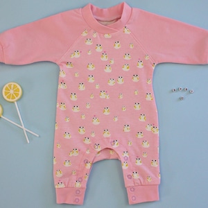 Könnte beinhalten: Rosaer Baby-Strampler mit einem Muster aus gelben Fröschen. Der Strampler hat lange Ärmel und einen Rundhalsausschnitt.