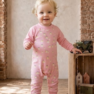 May include: A light pink long-sleeve romper featuring a repeating pattern of small, stylized yellow and white owl-like creatures. The garment has a crew neckline and ribbed cuffs at the wrists and ankles, paired with soft, light brown suede-like booties.