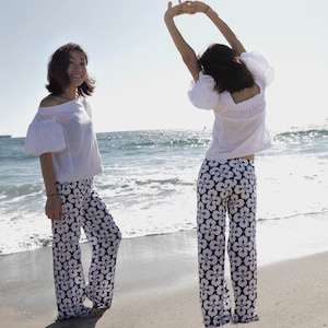 May include: Two women wearing white tops and black and white floral print pants. The women are standing on a sandy beach with the ocean in the background.
