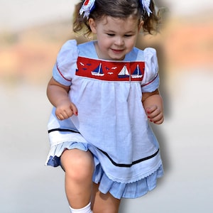 May include: A light blue and white nautical-themed dress with red accents. The dress features a red panel with sailboat and bird embroidery. The sleeves and hem are trimmed with red and black details. The dress is worn by a young child.