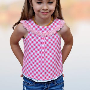 Könnte beinhalten: Ein junges Mädchen trägt ein ärmelloses Oberteil aus rosa und weißem Gingham mit einem rosa Blumenbesatz am Ausschnitt. Das Oberteil hat einen weißen Spitzenbesatz am Saum.