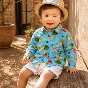 May include: A light blue long-sleeve button-down shirt featuring a repeating pattern of green frogs and red spotted mushrooms, paired with white shorts, a tan straw fedora hat with a light blue striped band, and brown leather boat shoes.
