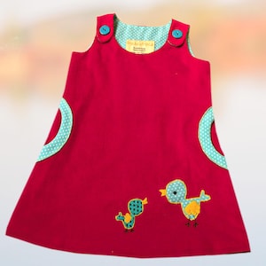 Pode incluir: Vestido vermelho sem mangas com detalhes em bolinhas turquesa e aplicações de pássaros. O vestido tem botões turquesa nas alças e bolsos de bolinhas turquesa. Duas aplicações de pássaros são costuradas na frente.