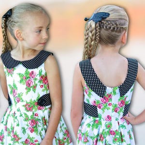 Könnte beinhalten: Zwei junge Mädchen tragen ein weißes Kleid mit einem schwarz-weißen Polka-Dot-Kragen und einem Blumendruck. Das Kleid hat ein schwarzes Band um die Taille. Ein Mädchen hat ihre Haare in einem Zopf mit einer Schmetterlingshaarspange. Das andere Mädchen hat ihre Haare in einem Pferdeschwanz.