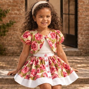 May include: A two-piece outfit consisting of a short-sleeved bolero jacket and a flared skirt, both featuring a vibrant pink rose floral pattern on a green background. The skirt has a white hem and a vertical row of pink buttons down the front.