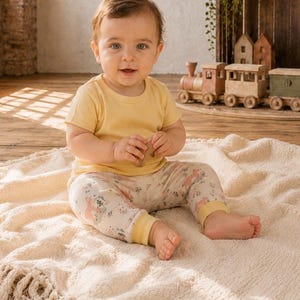 May include: A baby wearing a short-sleeved pale yellow t-shirt and cream-colored leggings featuring a delicate floral and animal print pattern. The leggings have solid yellow cuffs at the ankles, matching the color of the t-shirt worn by the baby.