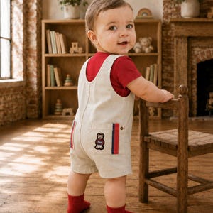 May include: A red short-sleeved t-shirt worn under beige cotton overalls. The overalls feature a back pocket with a small embroidered bear patch and vertical red and blue striped trim on the sides. The outfit is paired with solid red ankle socks.