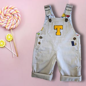Peut inclure: Salopette beige avec une lettre T en feutre jaune sur le devant. La salopette a des bretelles réglables et une fermeture à bouton.