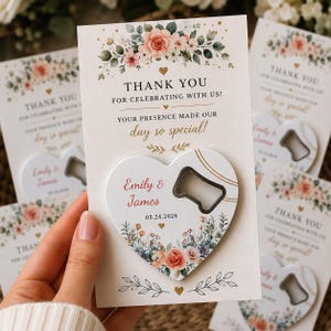 Pode incluir: Um abre-garrafas em forma de coração preso a um cartão retangular. O cartão apresenta ilustrações florais em pêssego e verde, com o texto: THANK YOU FOR CELEBRATING WITH US! YOUR PRESENCE MADE OUR day so special! O abre-garrafas exibe Emily & James 05.24.2026.