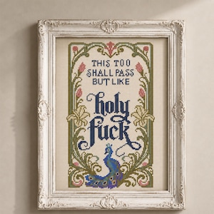 May include: A framed cross-stitch embroidery featuring the text "THIS TOO SHALL PASS BUT LIKE holy fuck" in dark blue lettering. The design is surrounded by a decorative border of green vines, pink flower buds, and a blue and purple peacock.