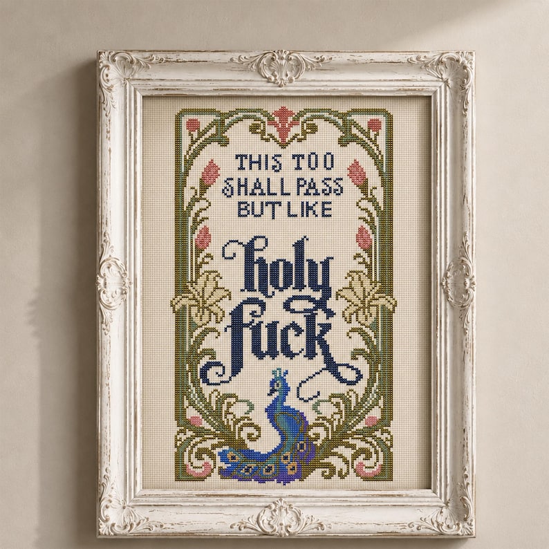 May include: A framed cross-stitch embroidery featuring the text "THIS TOO SHALL PASS BUT LIKE holy fuck" in dark blue lettering. The design is surrounded by a decorative border of green vines, pink flower buds, and a blue and purple peacock.
