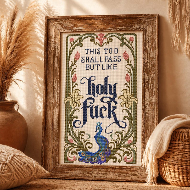 May include: A framed cross-stitch embroidery featuring the text 'THIS TOO SHALL PASS BUT LIKE holy fuck' in dark blue lettering. The design is surrounded by a decorative border of green vines, pink flower buds, and a blue peacock at the bottom.