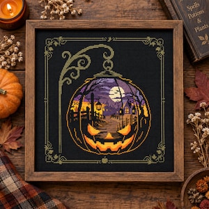 Puede incluir: Un bordado de punto de cruz enmarcado sobre tela negra que presenta una calabaza brillante con forma de farol colgante. El diseño muestra una escena nocturna con luna llena, árboles desnudos, una valla y una casa, todo dentro de un borde decorativo.