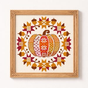 Puede incluir: Un bordado en punto de cruz enmarcado que presenta una calabaza central con variados patrones geométricos en tonos naranja, rosa y blanco, rodeada por una corona circular de hojas de otoño y bellotas estilizadas en amarillo, dorado y rojo oscuro.