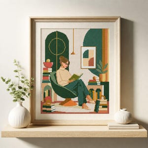 Puede incluir: Un bordado en punto de cruz enmarcado que representa a una persona sentada en una silla verde oscuro leyendo un libro. La escena incluye pilas de libros, una planta en maceta y un cuadro en tonos verde, beige, naranja y marrón en un marco de madera.