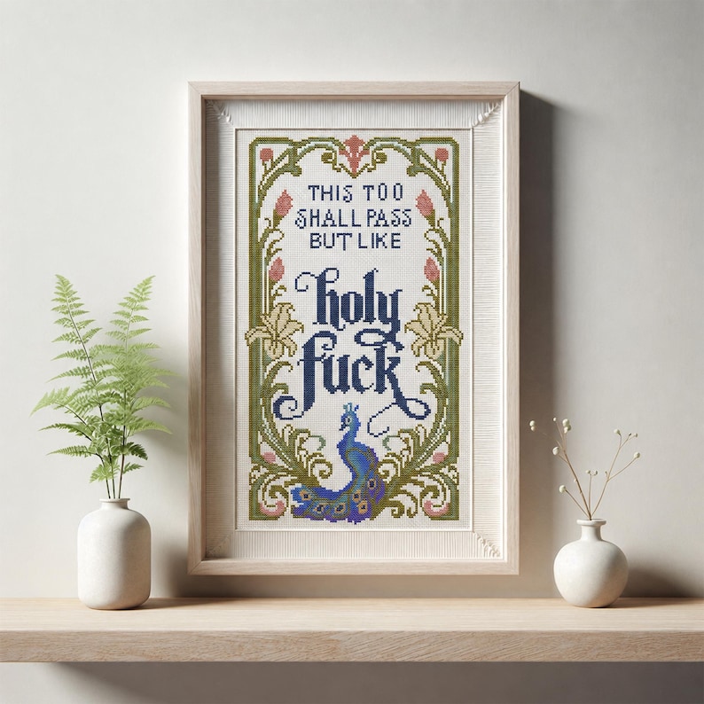May include: A framed cross-stitch embroidery featuring the text "THIS TOO SHALL PASS BUT LIKE holy fuck" in dark blue lettering. The design is surrounded by a decorative border of green vines, pink flower buds, and a blue peacock at the bottom.