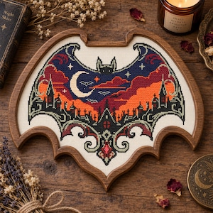May include: A cross-stitch embroidery piece framed in a dark wood bat-shaped frame. The design features a night scene with a large bat silhouette, a crescent moon, stars, and a dark castle landscape in shades of navy blue, orange, and red.