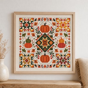 May include: A square cross-stitch embroidery in a light wood frame featuring a central geometric star pattern surrounded by orange pumpkins, stacked gourds, and autumn leaves in shades of green, yellow, red, and orange on a cream fabric background.